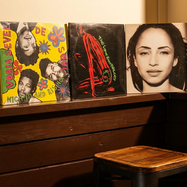 Three vinyl records, including Sade and A Tribe Called Quest, displayed on a wooden shelf above a stool.