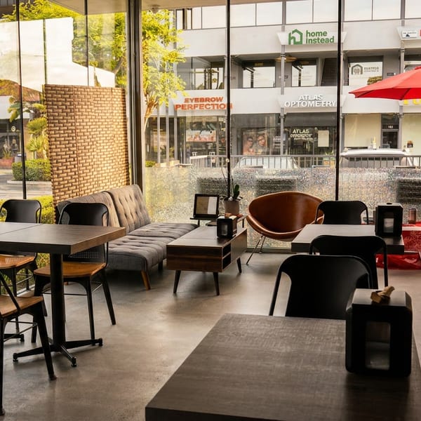 Restaurant interior with tables, chairs, and a sofa, looking out through large windows to a street.