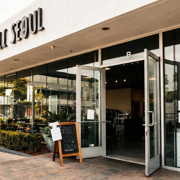 The exterior of Deli Seoul, a Korean deli, with an open glass door and a "Service Dogs Only" sign.