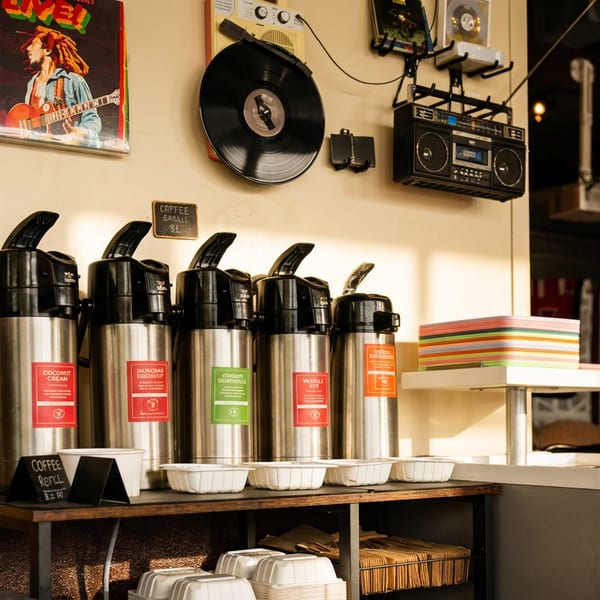 Coffee station with multiple dispensers, cups, and creamers, under a wall decorated with music memorabilia.