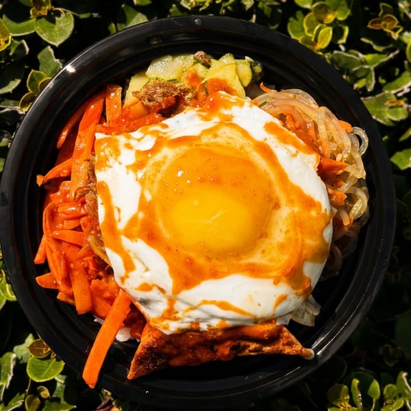 A black bowl of bibimbap with a fried egg, carrots, and other vegetables, on a green leafy background.