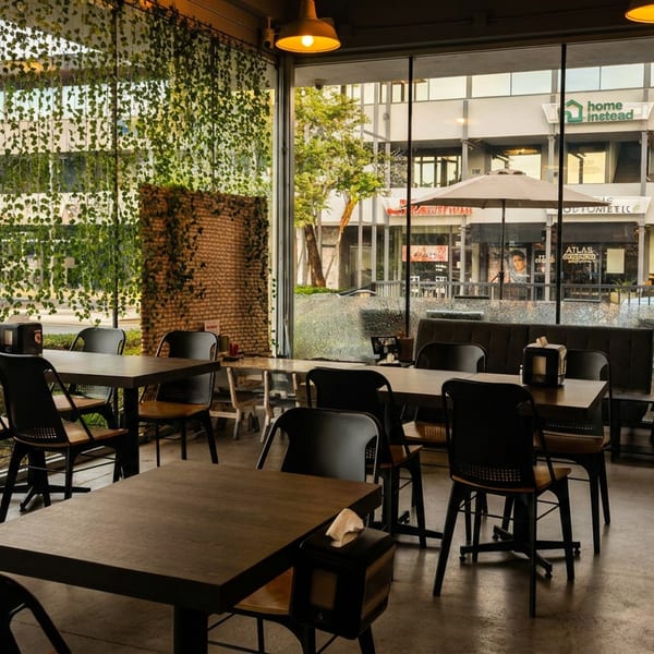 Restaurant interior with dark tables and chairs, large windows, and hanging green vines, looking out onto a street.
